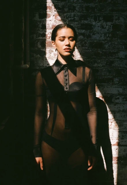 A young woman with her eyes closed stands in a beam of light against a brick wall. She's wearing a dark, sheer shirt with a collar.
