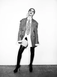 Model wearing an oversized grey blazer, white shirt, patterned tie, and thigh-high black boots against a plain white wall