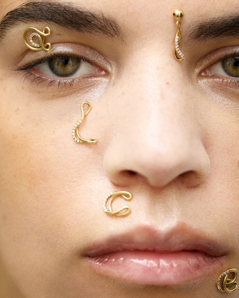 Close-up of a model wearing golden jewelry as piercings, including her nose, eyebrow, and forehead. The accessories match her tan skin.