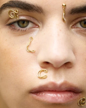 Close-up of a model wearing golden jewelry as piercings, including her nose, eyebrow, and forehead. The accessories match her tan skin.