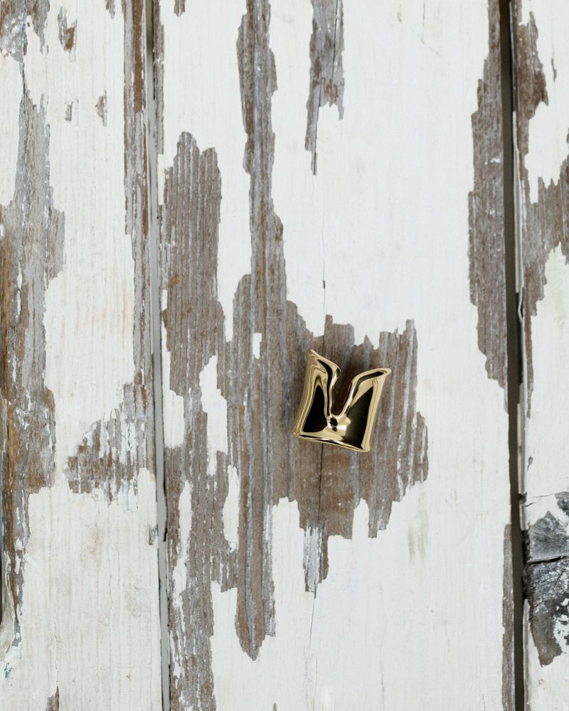 Elevated close-up of a gold-colored letter 'M' emblem resting on a weathered white wooden surface. The emblem is glossy, reflecting light.