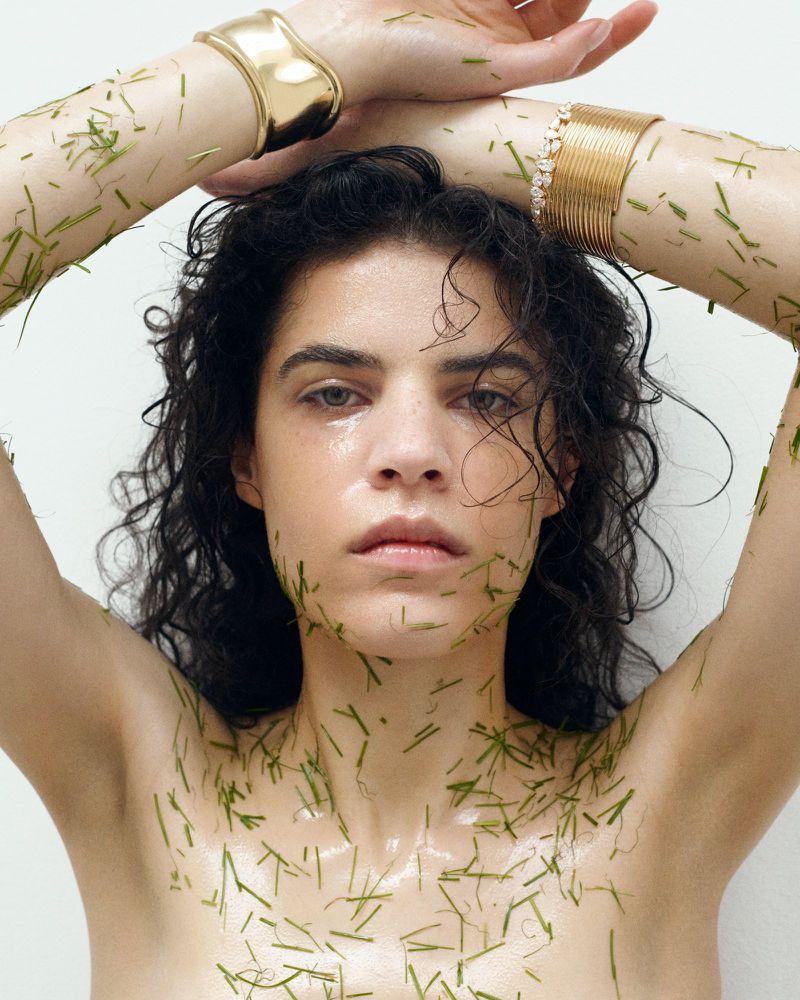 A young woman with wet dark curly hair poses with green grass blades on her bare skin and arms while wearing gold cuff bracelets.