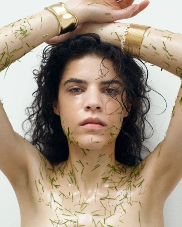 A young woman with wet dark curly hair poses with green grass blades on her bare skin and arms while wearing gold cuff bracelets.