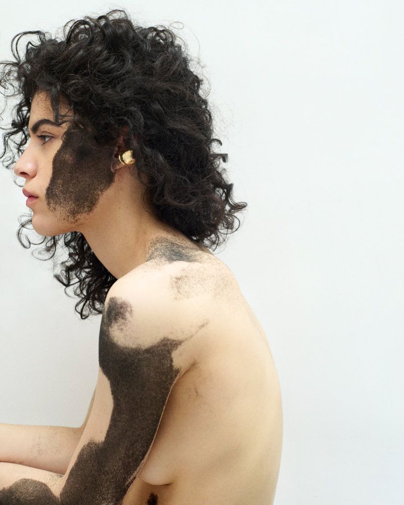 Profile view of a woman with curly hair and artistic black makeup on her face and body against a white background in a studio setting.