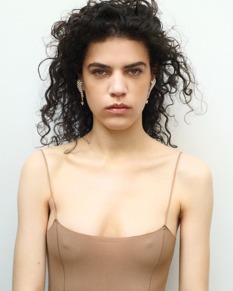 Close-up portrait of a young adult woman with curly dark hair wearing earrings, a light brown dress, and posing in front of a white wall.