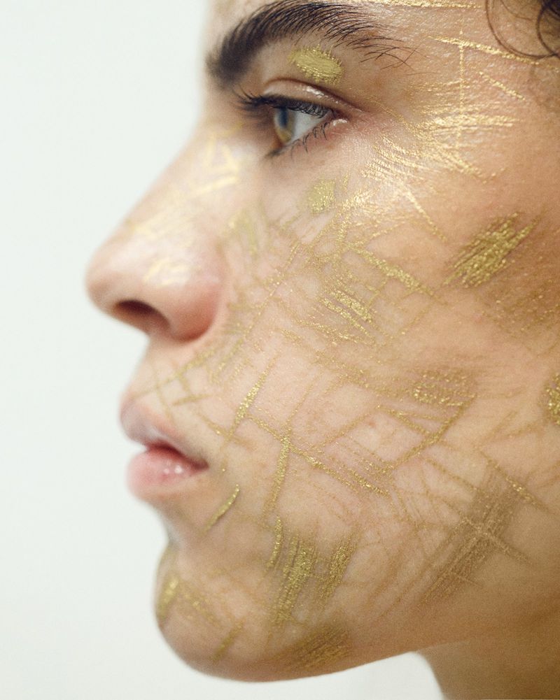 Adult female with gold face paint in profile, wearing beauty makeup against a white backdrop in a studio. Beauty and artistic fashion.