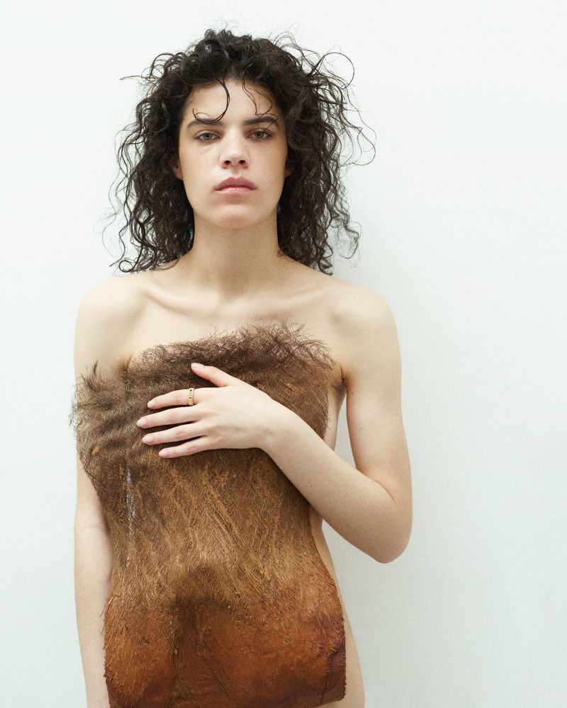 Striking portrait of a young woman in a studio, partially covered by a textured brown textile, against a stark white background.