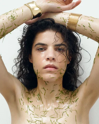 Model with wet dark hair and grass clippings on skin wearing gold cuffs against a white background