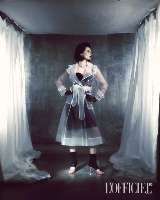 Elegant woman in a sheer trench coat posing indoors against a grey backdrop, framed by draped curtains. Fashion editorial shot.