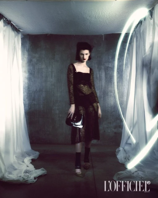 Elegant adult female fashion model strikes pose in a photo studio wearing a dark dress with a purse and high heels against a dark backdrop.