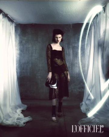 Elegant adult female fashion model strikes pose in a photo studio wearing a dark dress with a purse and high heels against a dark backdrop.