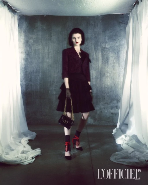 A fashion editorial shot of a model in a polka-dot jacket and tiered skirt holding a small chain purse in a studio setting.