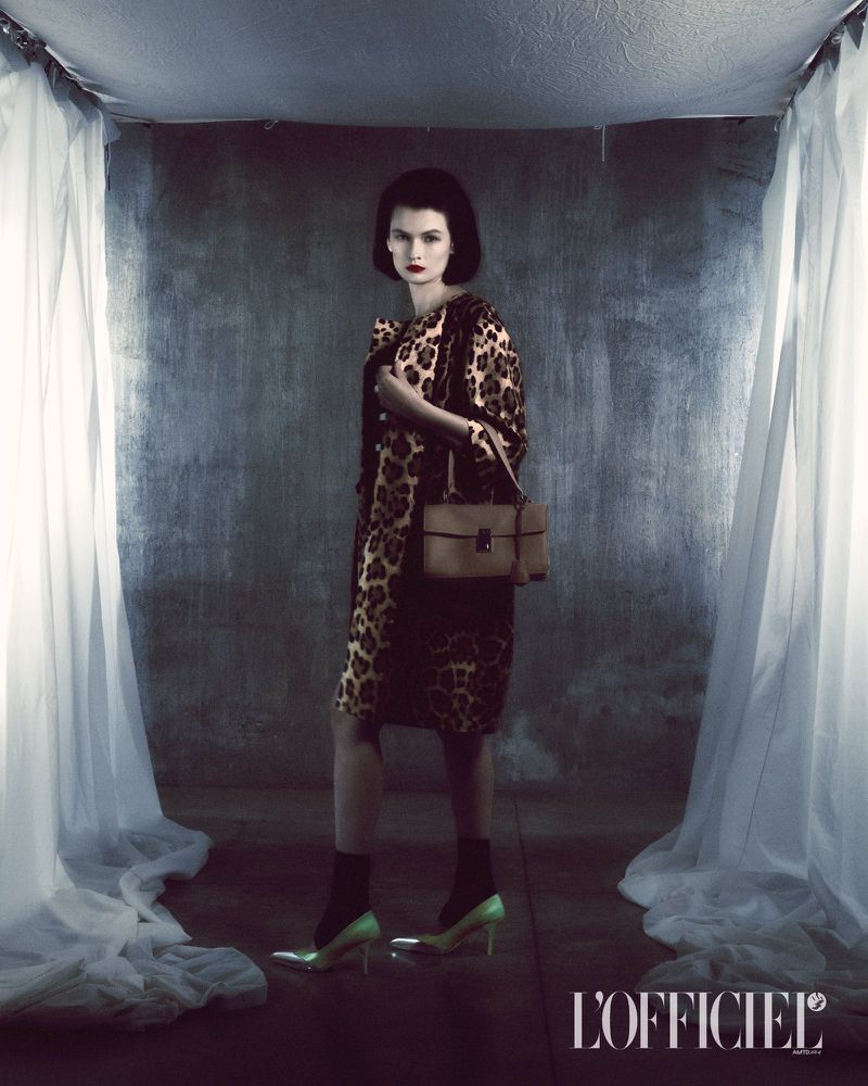 Elegant woman in leopard-print dress and coat accessorized with a handbag and high heels, set against a textured grey backdrop.