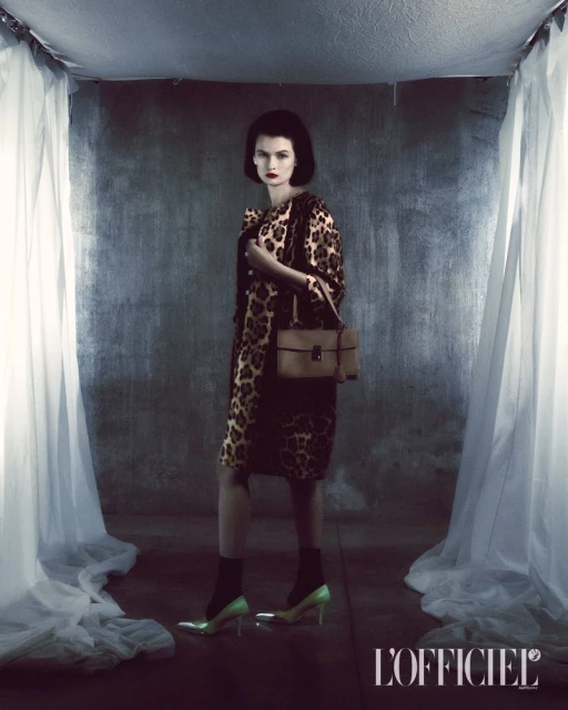 Elegant woman in leopard-print dress and coat accessorized with a handbag and high heels, set against a textured grey backdrop.