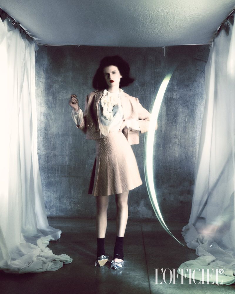 A fashionable woman in a pastel skirt suit stands in a minimalist setting with a glowing arch and sheer curtains for a magazine editorial.