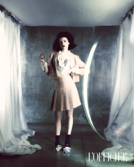 A fashionable woman in a pastel skirt suit stands in a minimalist setting with a glowing arch and sheer curtains for a magazine editorial.