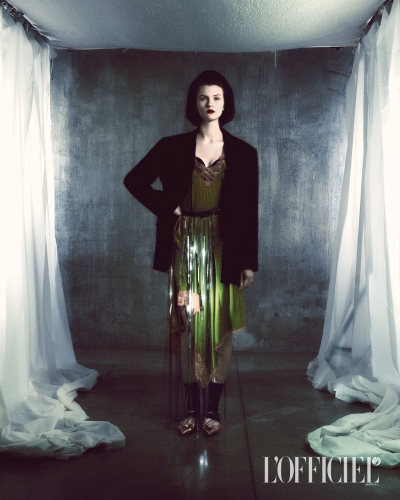 A stylish woman in a green dress and black blazer poses in a studio with draped white fabric and grey backdrop, creating an editorial fashion shot.