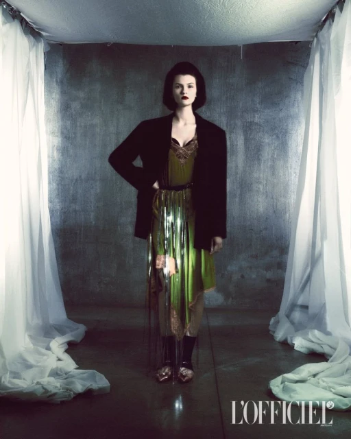 A stylish woman in a green dress and black blazer poses in a studio with draped white fabric and grey backdrop, creating an editorial fashion shot.