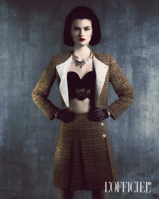 A fashionable woman wearing a tweed skirt and jacket with a black lace bralette stands against a textured grey backdrop in a studio shot.