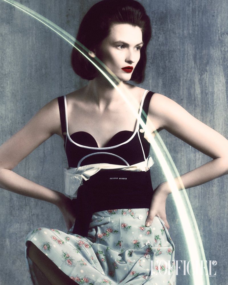 Elegant woman in black top and floral pants posing with light effects against a grey backdrop in a fashion editorial style.