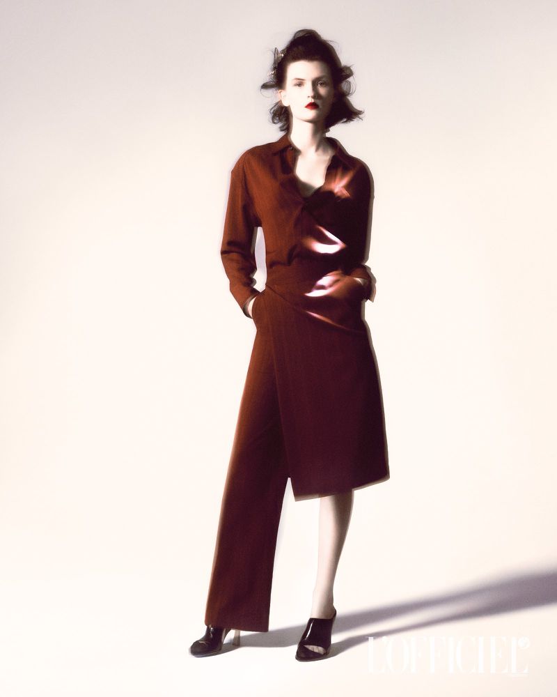 A fashionable brunette woman stands in a photo studio wearing a chic, brick-red button-down outfit with a high-fashion wrap skirt.