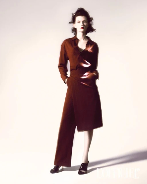 A fashionable brunette woman stands in a photo studio wearing a chic, brick-red button-down outfit with a high-fashion wrap skirt.