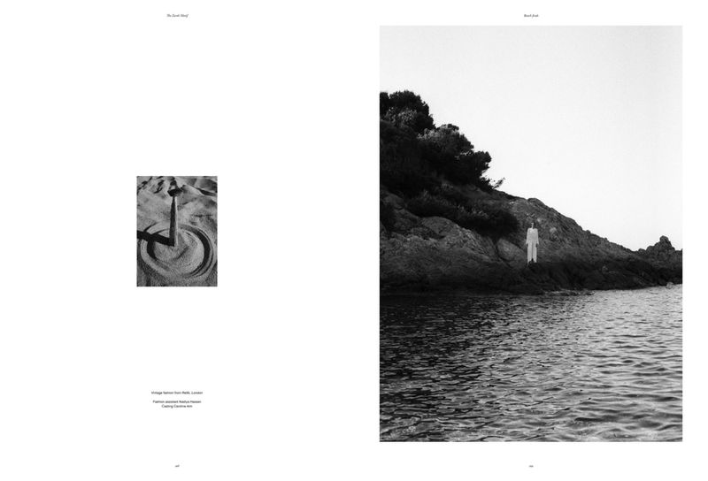 Black and white photo of an artful sand circle with a stick, contrasting with a distant figure by a rocky shoreline.