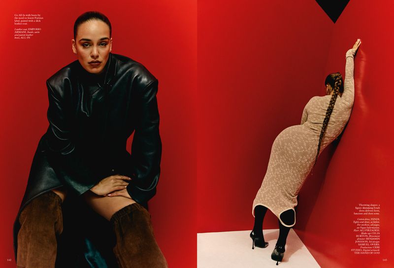 Two adult women in a studio setting. One is seated in a black leather coat and brown boots, the other is standing against a red wall.