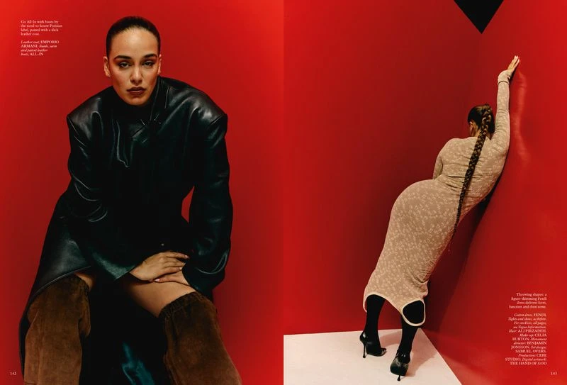 Two adult women in a studio setting. One is seated in a black leather coat and brown boots, the other is standing against a red wall.