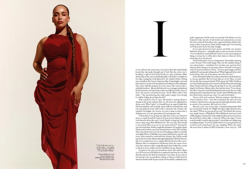 Studio portrait of Jorja Smith wearing a red dress with a braided ponytail posing against a bright background, fashion editorial-style shot.