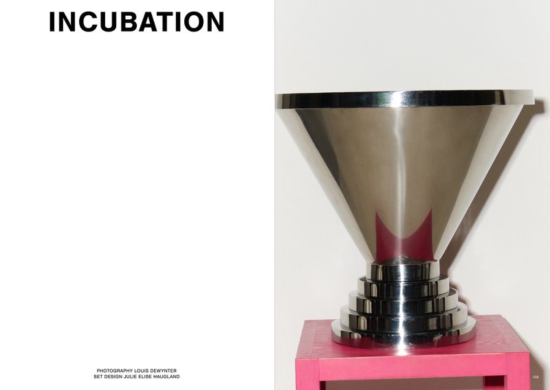 Studio shot of a polished metal art object with a cone and stepped base, sitting on a pink table against a white background.