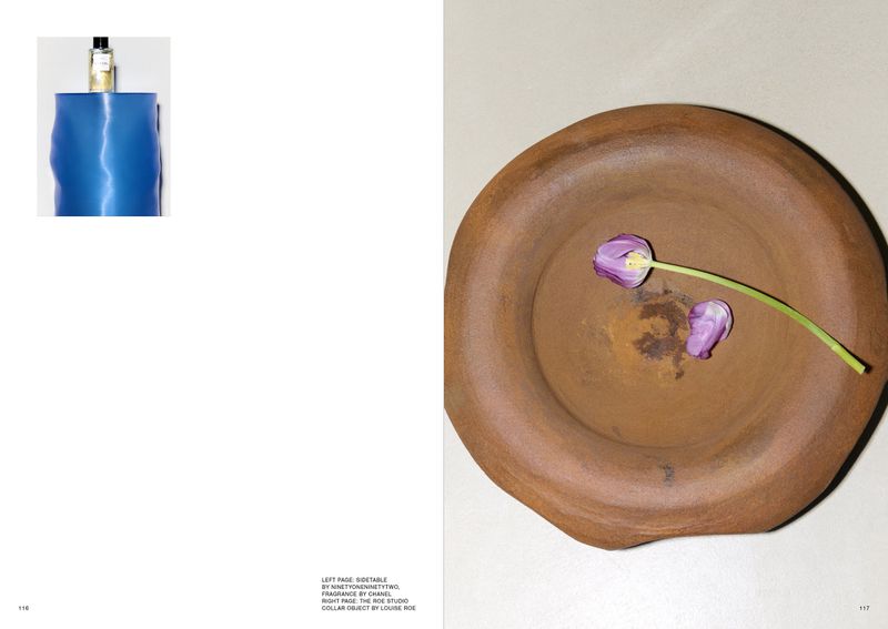 Still life photo features a blue perfume bottle and a rustic clay bowl with two purple tulip blossoms inside, set against a cream background.
