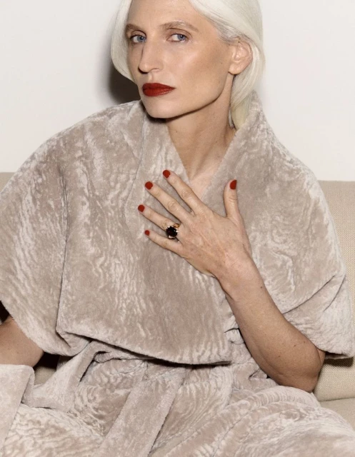 Elegant older woman with silver hair, red lipstick, and a statement ring, posing in a light-beige furry coat against a simple backdrop.