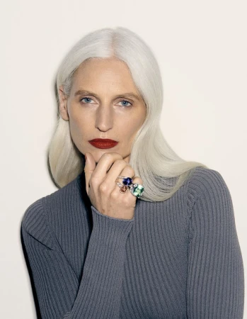 Close-up studio portrait of an adult woman with silver hair and red lipstick, wearing a gray turtleneck and three statement rings.