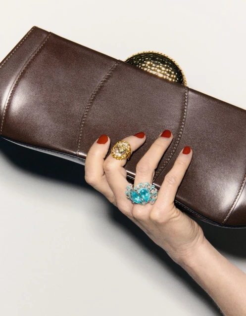 Hand holds brown leather clutch, adorned with gold and blue gem rings, against a plain background in a fashion editorial.
