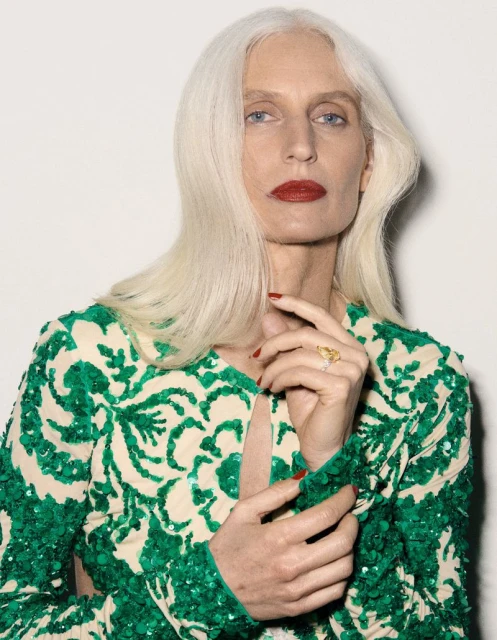 Close-up of an adult woman with long white hair, wearing a green and white floral patterned dress, posing against a white backdrop.