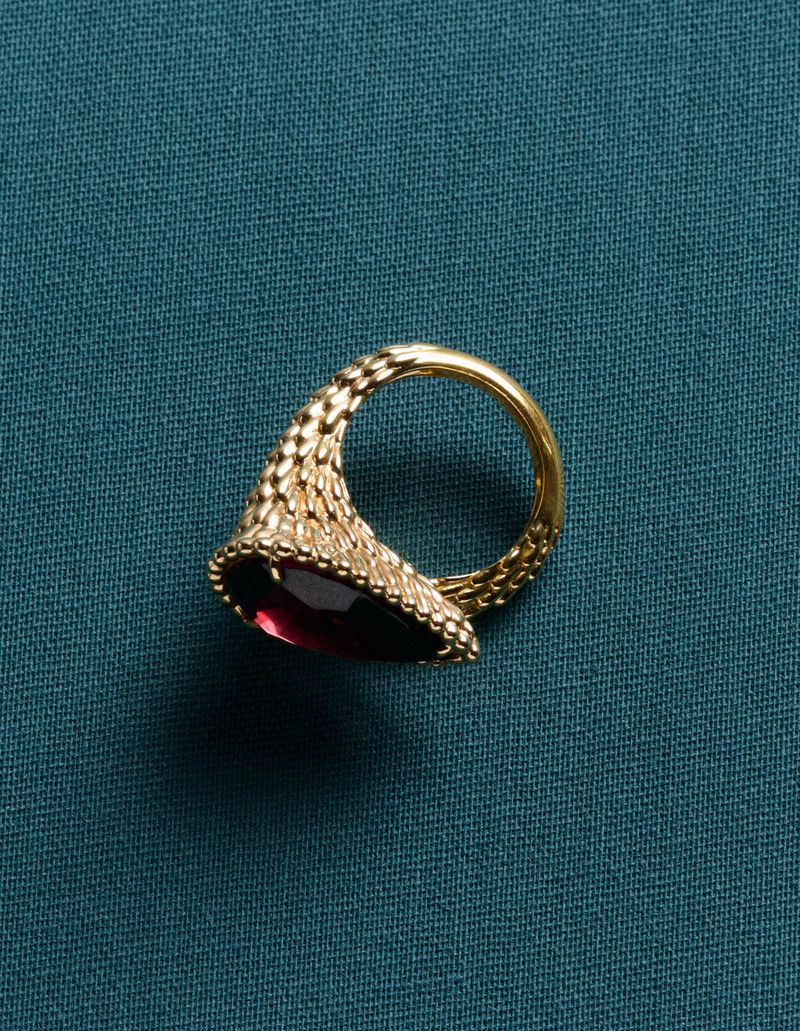Detailed view of an ornate gold ring featuring a pear-shaped red gemstone, set against a textured teal fabric backdrop.