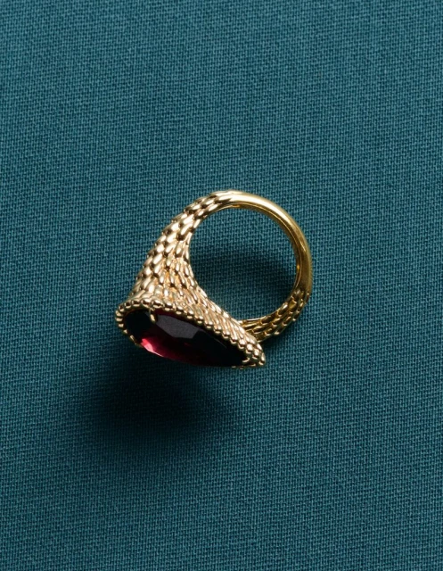 Detailed view of an ornate gold ring featuring a pear-shaped red gemstone, set against a textured teal fabric backdrop.