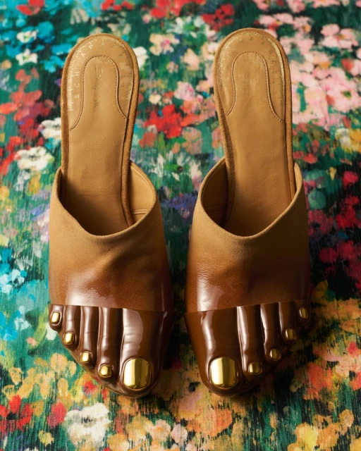 Overhead studio shot of two tan leather mule shoes with toes crafted in a darker tone with gold painted toenails on a floral background.