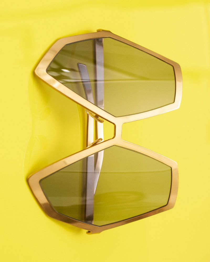 Geometric aviator sunglasses with gold frames and green lenses on bright yellow surface. Fashion eyewear product photography.