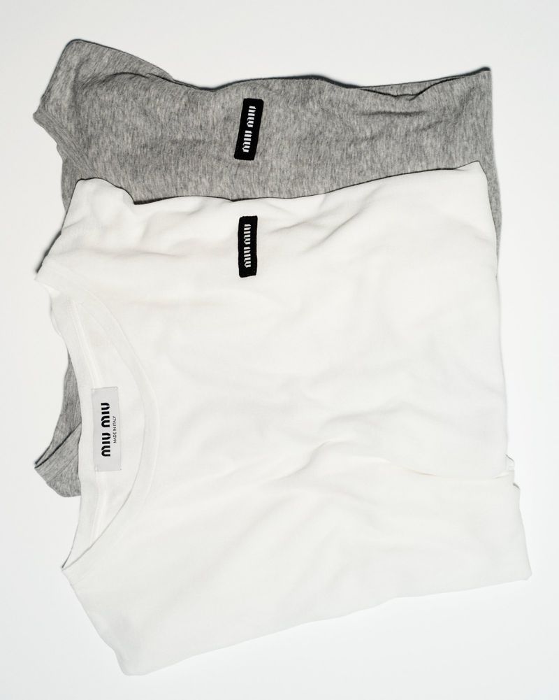 Gray and white shirts stacked in a studio shot, featuring the Miu Miu brand label. Simple studio lighting, minimal styling.