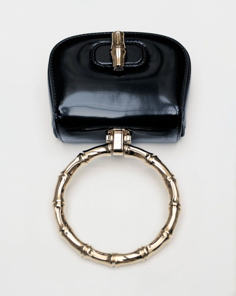 Shiny black handbag with a circular gold bamboo handle on a white background, showcasing luxury fashion and modern accessory design.