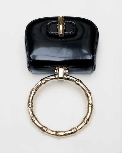 Shiny black handbag with a circular gold bamboo handle on a white background, showcasing luxury fashion and modern accessory design.