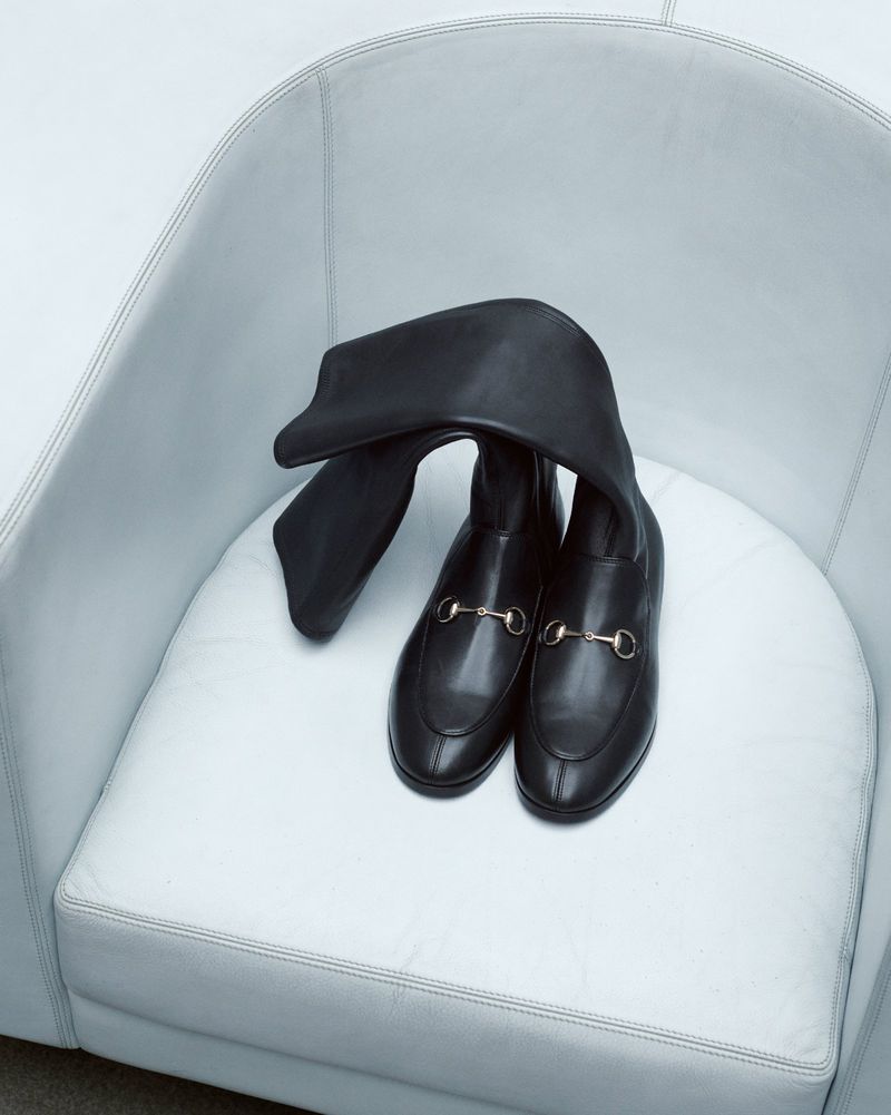Black leather heeled boots with gold hardware sit in a white round chair, product shot, minimal composition, fashion advertisement.