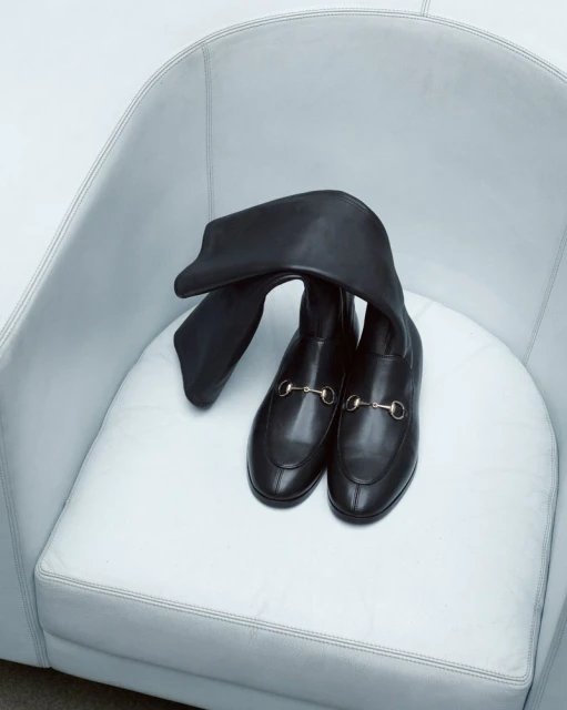 Black leather heeled boots with gold hardware sit in a white round chair, product shot, minimal composition, fashion advertisement.
