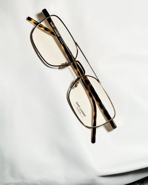 Stylish Saint Laurent eyeglasses with a thin gold frame and tortoiseshell arms are displayed on a smooth white surface.