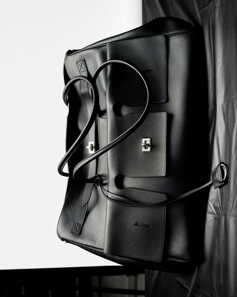 Black leather tote bag with silver hardware, photographed in a studio setting against a white and black draped backdrop.