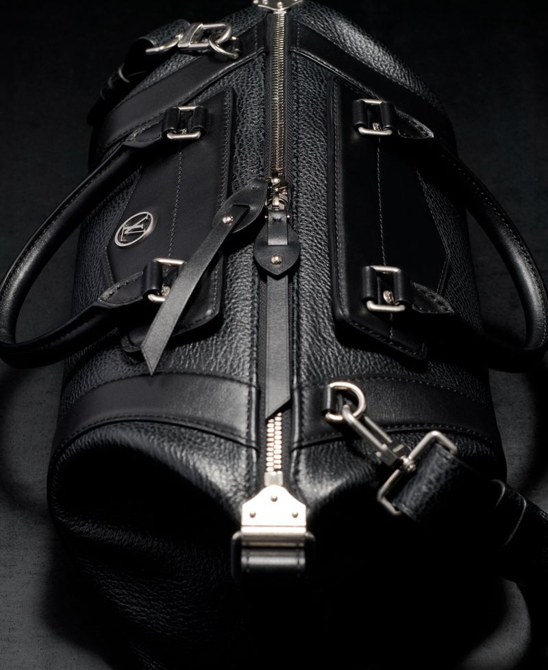 Close-up shot of a stylish black leather duffel bag featuring a silver zipper, accents, and a detachable shoulder strap on a dark background.