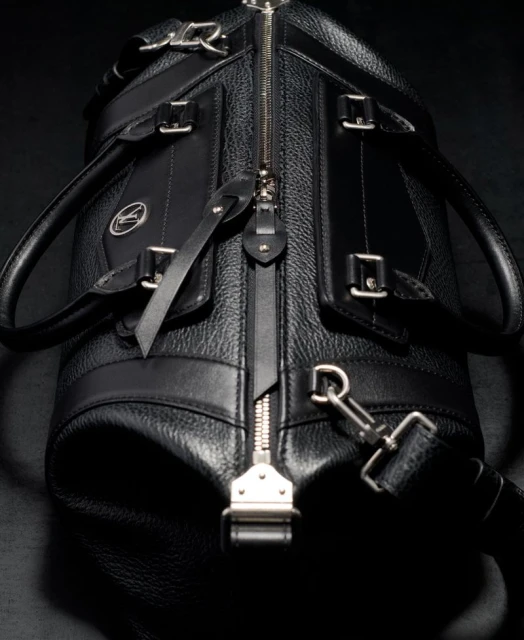 Close-up shot of a stylish black leather duffel bag featuring a silver zipper, accents, and a detachable shoulder strap on a dark background.