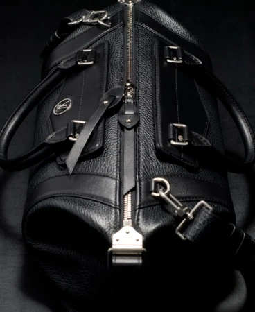 Close-up shot of a stylish black leather duffel bag featuring a silver zipper, accents, and a detachable shoulder strap on a dark background.